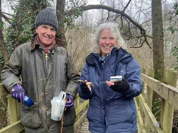 Dot and Mike Isgrove smiling. Dot is hodling a notebook and pen, Mike is holding a jug and testing instruments.