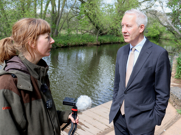 Fiona holds a microphone and is talking with Gideon Amos next to bathing place on the River Tone in French Weir Park in Taunton