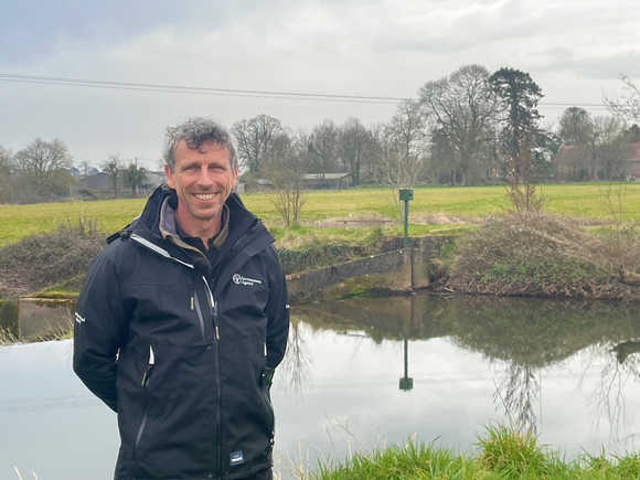 Oliver Hill smiling, in waterproofs, standing in front of the weir and fish pass at Bishop's Hull