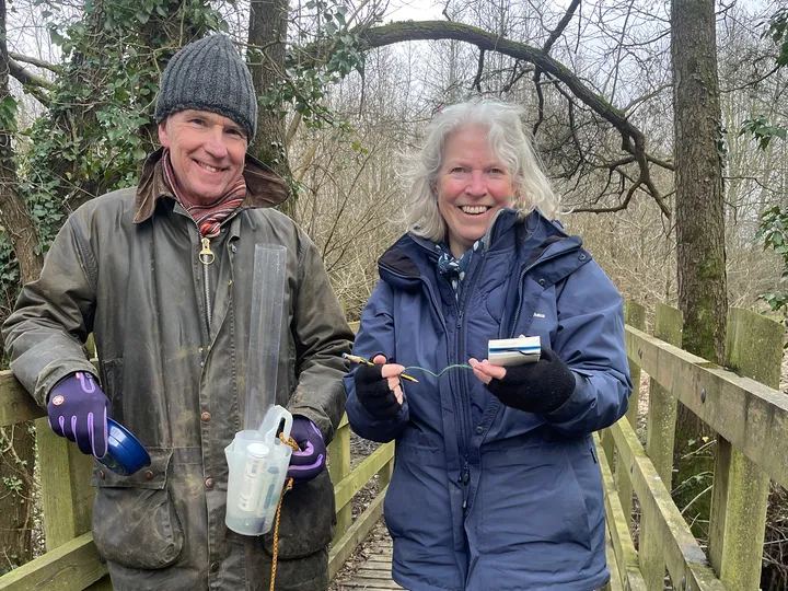 Dot and Mike Isgrove smiling. Dot is hodling a notebook and pen, Mike is holding a jug and testing instruments.