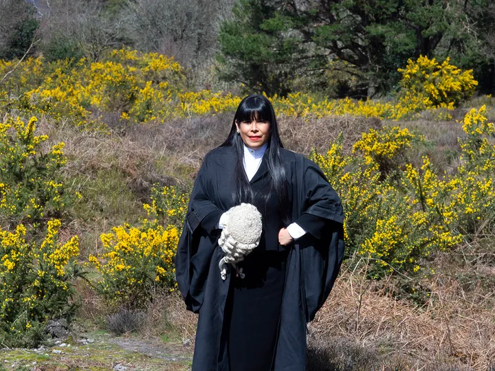 Monica Feria Tinta in her barrister's robes stands in a wild area of gorse and bracken